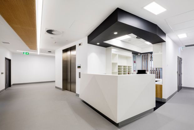 A bright, modern commercial reception area with a white angular reception desk, integrated shelving and workstations behind it, stainless-steel lift doors, grey flooring, and a bold black ceiling bulkhead with recessed lighting. The space has clean white walls, grey doors, and a suspended timber-toned acoustic ceiling feature to the left.