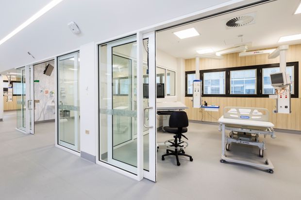 Bright, modern clinical hospital interior showing a patient room or treatment bay with a wheeled hospital bed, medical equipment, observation windows, glass-partitioned rooms, pale flooring, and light timber wall panelling. The space is clean, functional, and highly illuminated with recessed ceiling lights.