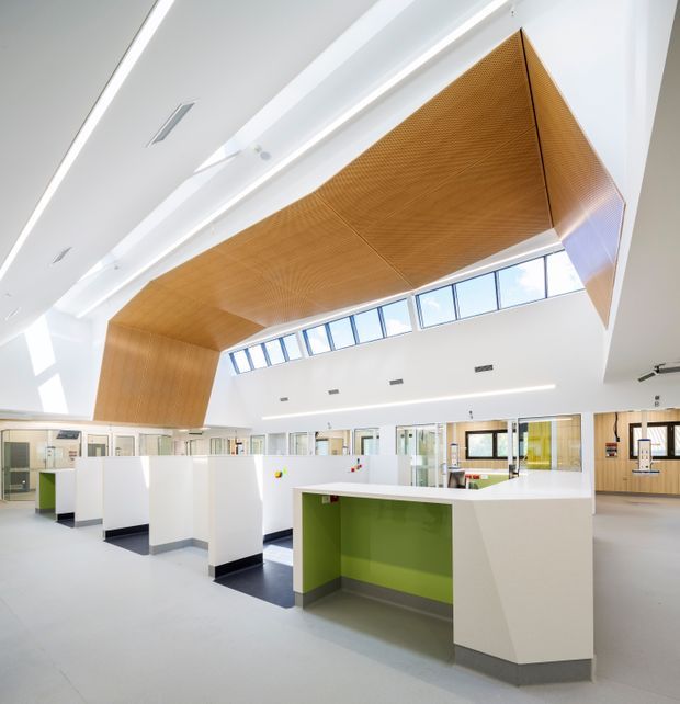 Bright contemporary commercial interior, likely a reception or collaborative workspace, with white service counters and partitions, a vivid green recessed counter face, pale flooring, extensive clerestory glazing, and a dramatic angular perforated timber ceiling feature. The space is open, highly illuminated, and institutional or office-like.
