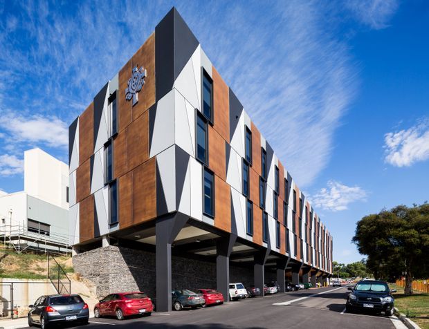 Exterior street-level view of a contemporary multi-storey commercial or institutional building elevated on dark structural columns, with a long repetitive facade composed of white angular panels, dark grey fins, and warm timber-look cladding. Cars and a driveway occupy the foreground, with blue sky and landscaping surrounding the building.