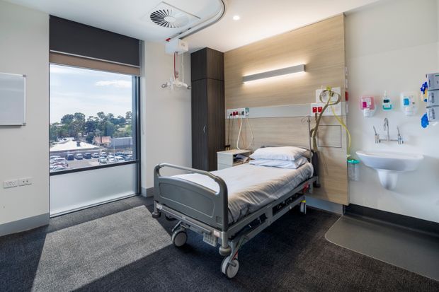 Bright modern hospital patient room with an adjustable medical bed, integrated bedside services and medical outlets, wall-mounted sink, dark carpet flooring, timber-look feature wall, and large window with roller blind. The room has a clean, functional healthcare fit-out and accessible circulation space.