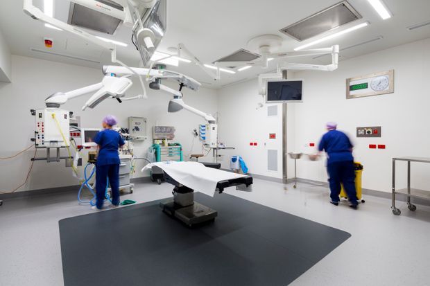 Bright, sterile hospital operating theatre with a central surgical table, multiple overhead surgical lights and equipment arms, wall-mounted medical devices and monitors, and two medical staff in blue scrubs. The room has white hygienic wall and ceiling finishes, pale flooring, and a large dark grey floor zone beneath the operating table.