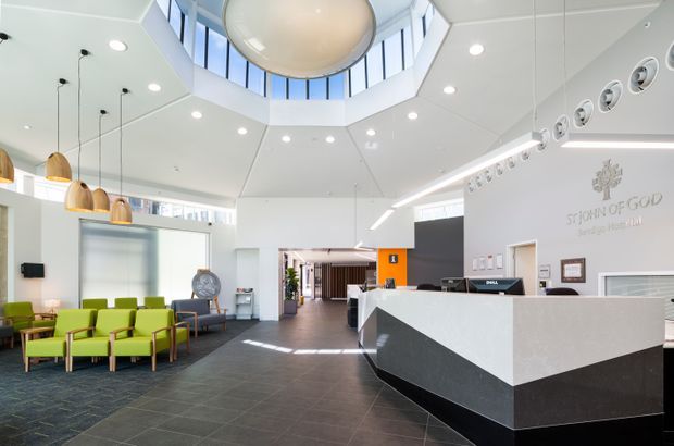 Bright contemporary reception lobby with a large angular white reception desk featuring a dark charcoal base, lime-green lounge chairs, dark tiled flooring, pendant lights, recessed lighting, and extensive clerestory glazing. A dramatic faceted ceiling rises to a central circular skylight or roof lantern, while the wall behind reception includes signage and a row of circular decorative or acoustic elements.
