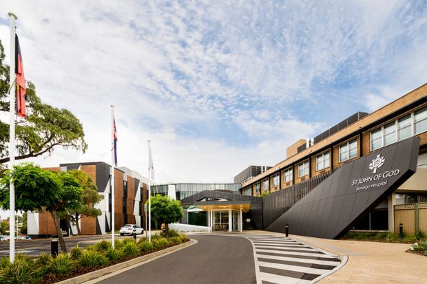 Exterior view of a contemporary multi-storey institutional building, identified by signage as Sydney Adventist Hospital's 'Sydney Adventist' facility. The image shows a broad paved drop-off driveway, striped pedestrian crossing, landscaped planting beds, flagpoles, and a glazed entrance canopy. The facade combines dark metal cladding, warm timber-toned panels, extensive horizontal windows, and a dramatic angular projecting entry feature.