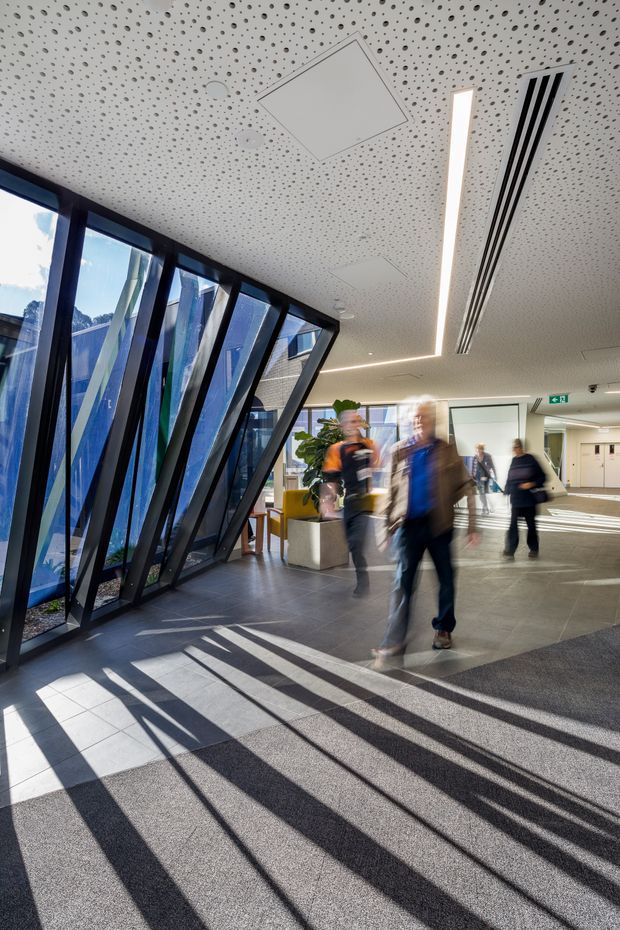 A bright contemporary commercial building lobby or circulation space with a high perforated acoustic ceiling, linear recessed lighting, dark tiled flooring, and dramatic angled floor-to-ceiling glazing. Sunlight casts long geometric shadows across the carpeted and tiled floor, while blurred pedestrians move through the open interior. A large colourful abstract glazed feature is visible along the left side.