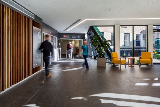 A bright contemporary institutional lobby or reception area with a spacious tiled floor, timber vertical-slat feature wall, dark grey walls, directional signage, floor-to-ceiling glazing, and a small seating arrangement of mustard-yellow armchairs around timber side tables. People are walking through the space, and a large indoor plant sits beside an angled structural or glazed feature.