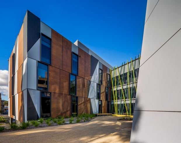 Exterior architectural photograph of a contemporary multi-storey building with a sharply articulated facade. The envelope combines dark and silver-grey panels with warm timber-toned cladding, tall narrow windows, and a prominent screened section of angled green and yellow structural members. A paved pedestrian forecourt and landscaped planting sit around the building, while another light-coloured facade frames the right side.