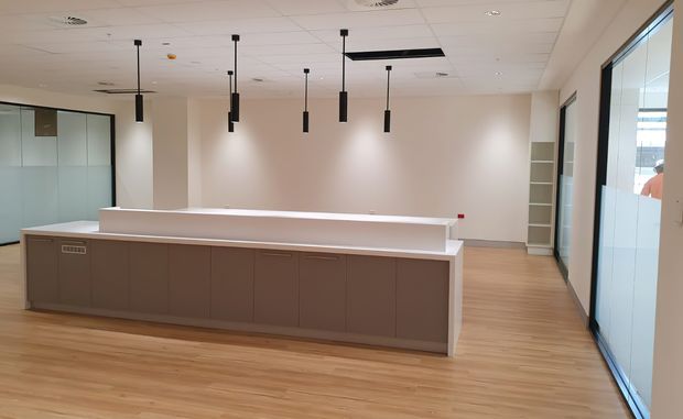 A contemporary commercial reception or service area with a long taupe built-in counter, pale timber-look flooring, white walls and ceiling, black suspended pendant lights, glazed office partitions, and full-height glass doors.