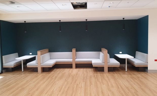 A modern commercial lounge or waiting area with built-in upholstered bench seating arranged along a deep blue accent wall. The space features pale timber-look flooring, light wood and white seating components, slim black pendant lights, and a suspended acoustic tile ceiling with recessed lighting.