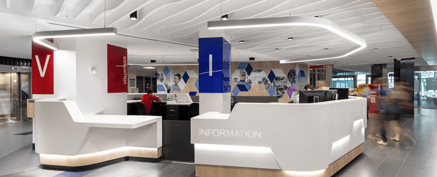 A bright, contemporary commercial reception and information desk in a large public interior. White angular reception counters with integrated linear lighting sit beneath a sculptural white slatted ceiling with recessed spotlights and illuminated geometric ceiling fixtures. Blue, red, black and white branding elements, geometric wall graphics, computers and staff workstations are visible behind the desks. The space has dark grey tiled flooring, glass entry doors, lift doors and a polished, high-traffic institutional or corporate atmosphere.