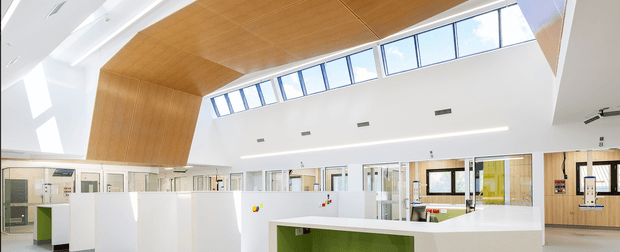 Bright contemporary institutional interior, likely a school or commercial learning space, with an open-plan layout, white partition walls, large clerestory glazing, and extensive warm timber wall and ceiling lining. A prominent white reception or work counter with a green inset panel occupies the foreground. The high ceiling and abundant daylight create an airy, clean environment.
