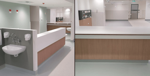 A split view of a clean, contemporary commercial healthcare or public facility interior. The left side shows a wall-mounted white basin with exposed plumbing, soap and paper dispensers, and a pale green-gray wall. Behind it is a reception or service counter with a white solid-surface top and light timber-look fixed cabinetry. The right side focuses on the same counter, with a wood-grain front, white benchtop, pale flooring, and a simple corridor with doors and signage in the background.