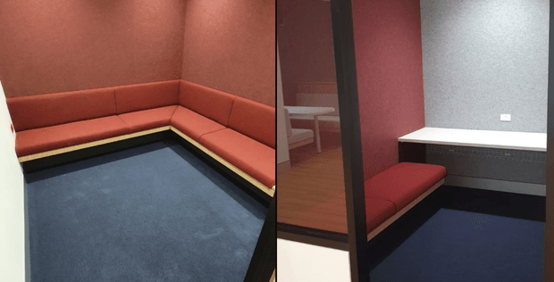 A compact, enclosed meeting or lounge nook with a built-in L-shaped upholstered bench in deep red, set against red walls. The space has dark blue carpet, a contrasting light timber or pale trim beneath the seating, and a dark-framed glazed partition or door revealing a small adjoining area with a table. The design is functional and contemporary with strong, muted colour blocking.