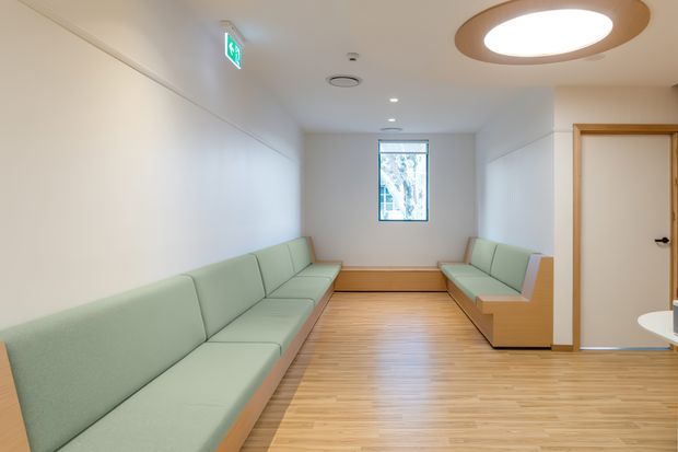 A bright, narrow institutional or commercial waiting area with long built-in pale green upholstered benches along both walls. The room has a warm light timber floor, plain white walls, a small rear window, a timber-framed door, recessed ceiling lights, and a large circular flush-mounted ceiling fixture with a wood-toned surround. An illuminated exit sign is visible near the ceiling.