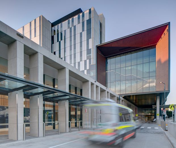 Urban exterior view of a contemporary multi-storey commercial or institutional building complex. The composition features pale concrete columns and a glazed canopy in the foreground, a tall facade clad in alternating light panels and dark glazing, and a prominent projecting volume with a deep red metal roof and large glass curtain wall. A blurred city bus is passing through the covered ground-level access area.