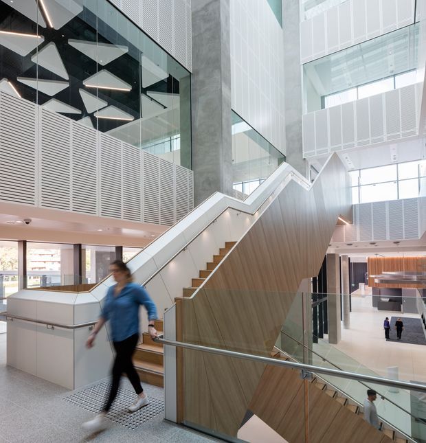 A bright, multi-level contemporary commercial interior focused on a sculptural stairway. The stair has warm wood-clad sides, pale treads, glass balustrades, and integrated linear lighting. Surrounding atrium spaces feature extensive glazing, perforated or slatted acoustic wall panels, exposed concrete, white architectural surfaces, and suspended geometric pendant lights. Several people provide scale, with additional floors and circulation spaces visible in the background.