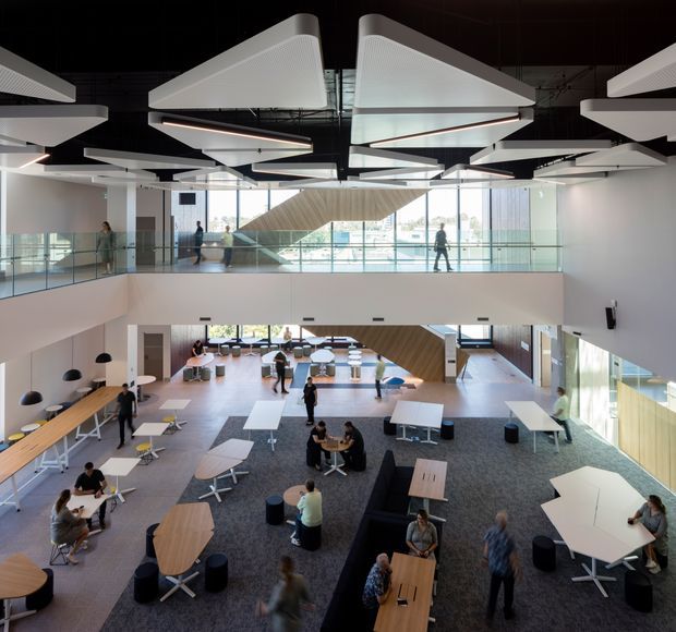 A large, contemporary multi-level commercial interior viewed from an upper landing, with an open-plan communal workspace or collaboration lounge below. The space features extensive glazing, glass balustrades, timber-clad stair and wall elements, grey carpet, light timber floors, movable tables and stools, and dramatic suspended white geometric acoustic ceiling panels. People are using the flexible work and meeting areas.