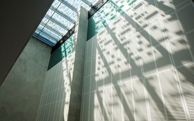 Upward-looking view of a tall, enclosed architectural atrium or internal void. A large glazed skylight admits daylight, casting strong geometric shadows across pale perforated or louvred wall panels. Exposed concrete surfaces and dark teal accent wall areas create a stark, contemporary institutional or commercial interior.