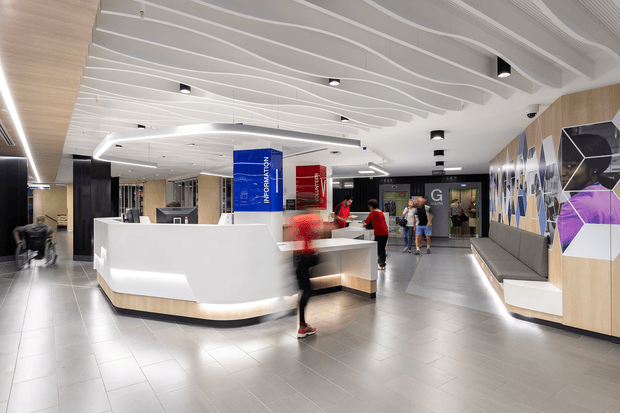 A bright, contemporary commercial reception and lobby area with a large curved white reception desk, integrated warm wood base, grey tiled flooring, black structural columns, and a sculptural white ribbed ceiling with recessed lighting. The right wall features built-in upholstered bench seating and a bold geometric mural in purple, blue, black, and white. Several people are present around the reception and entry area.