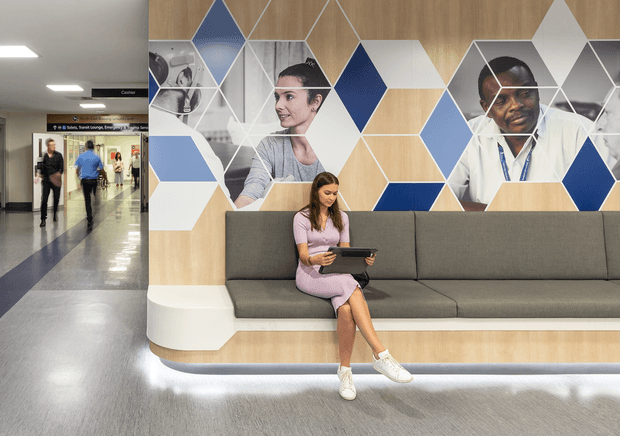 A contemporary healthcare or commercial reception waiting area with a long built-in charcoal upholstered bench, pale timber wall panelling, and a large geometric feature mural incorporating blue, white, and grey hexagonal forms with photographic portraits. A visitor sits using a tablet, while a bright corridor and directional signage extend to the left. The space has polished flooring, integrated under-seat lighting, and a clean, professional atmosphere.