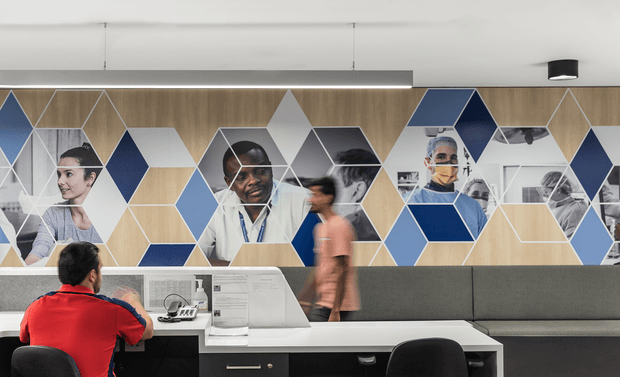 A contemporary commercial reception or consultation area with a long built-in desk, grey upholstered bench seating, black office chairs, and a large geometric wall mural featuring blue, beige, white, and grey diamond shapes overlaid with photographic portraits of medical professionals and patients. The space has a clean white ceiling, linear lighting, and dark built-in cabinetry beneath the desk. A seated person is using the workstation while another person walks past.