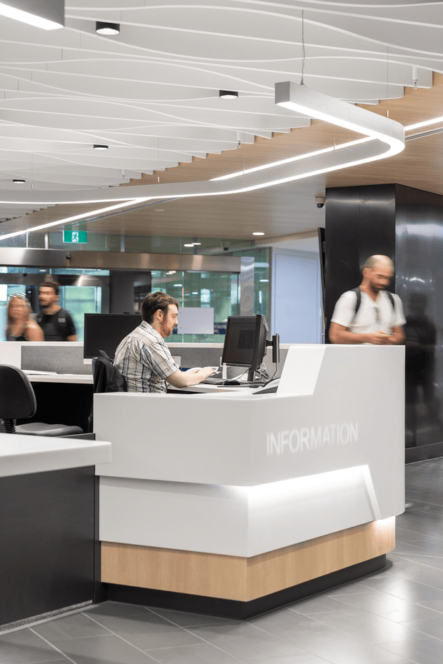 A modern commercial information or reception desk in a bright public interior. The white angular counter has integrated illuminated detailing and the word “INFORMATION” on its front, with a timber-look base and computer workstations. The space features a dark tiled floor, black feature wall, extensive glazing, timber ceiling panels, and a sculptural linear LED lighting installation with curved and wave-like elements. People are present in the background and at the desk.