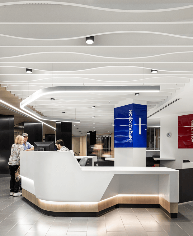 A bright contemporary commercial reception area with a sculptural white reception desk featuring illuminated under-counter joinery and light timber-toned detailing. The space has dark wall panels, grey tiled flooring, extensive glazing, blue and red information signage, and a distinctive ceiling with layered white curved linear elements and integrated lighting. Staff and visitors are present at the reception counter.