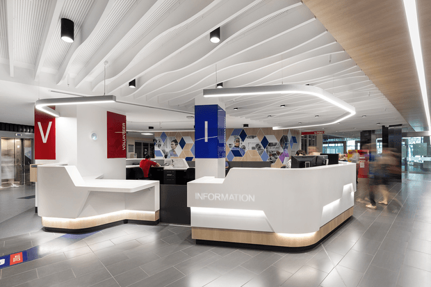 A bright contemporary commercial reception and information area, likely in a large public or corporate building. Two sculptural white reception desks with illuminated recessed bases dominate the foreground, with black workstations behind them. The space features a highly articulated white ribbed ceiling with suspended linear lighting, grey tiled flooring, a timber-toned ceiling inset and desk plinths, and bold red and blue branded feature panels. A geometric blue, grey and white wall graphic separates the reception area from the open circulation space.