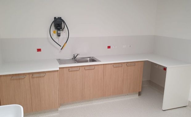 A clean, functional kitchenette or utility room with a long white countertop, stainless-steel sink and tap, light wood-look base cabinetry, white walls, and a compact under-counter appliance at the right. Multiple electrical outlets and exposed wall-mounted plumbing or utility connections are visible.
