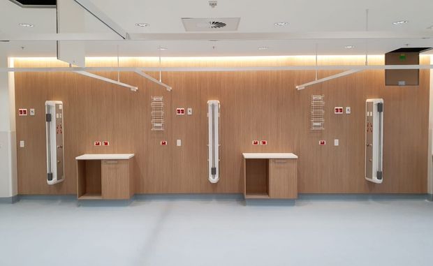A clean, modern institutional interior with a symmetrical timber-look wall panelled with medical equipment, wall-mounted fixtures, safety signage, and compact built-in timber counters. The pale resilient floor, recessed ceiling lights, linear concealed lighting, and exposed services suggest a hospital or clinical treatment area.