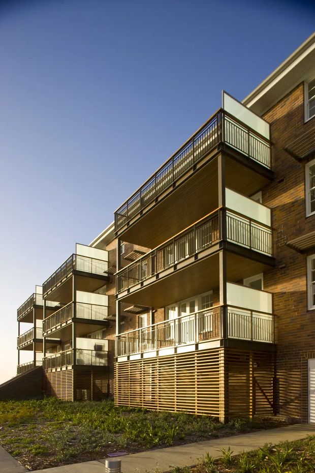Exterior view of a multi-storey brick apartment building with stacked projecting balconies, dark metal balustrades, translucent privacy screens, and timber-look horizontal screening at the lower levels. A paved walkway and landscaped planting sit in the foreground under clear daylight.