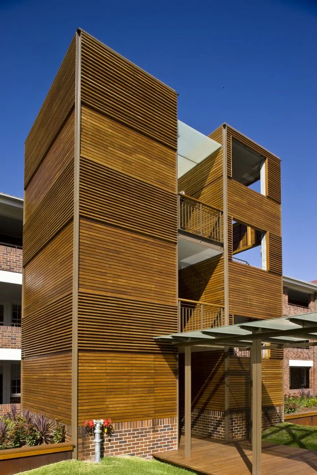 A tall contemporary multi-storey residential building or extension clad in warm horizontal timber slats, with projecting box-like volumes, open balconies, external stairs, and a translucent covered entry or walkway. Brickwork is visible at the base and on adjoining structures. The building is photographed from outside against a clear blue sky, with landscaped garden areas and a pergola-like canopy in the foreground.