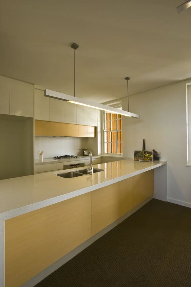 A contemporary galley-style kitchen with a large island and integrated double sink. The space features pale timber-look cabinetry, white upper cabinets, a light engineered stone benchtop, stainless steel appliances, a white splashback, and minimalist linear pendant lighting. A tall window with vertical timber screening brings warm natural light into the room, while dark carpet grounds the space.