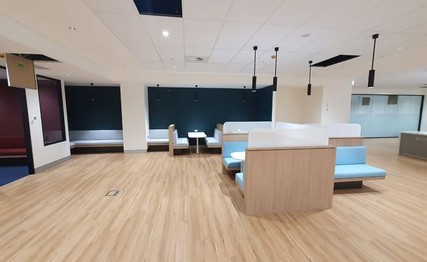 A bright contemporary commercial interior, likely a waiting or reception area, with pale timber-look flooring, built-in booth seating in light blue and white upholstery, pale wood privacy panels, dark feature walls, suspended black pendant lights, and a suspended ceiling with recessed services. Glazed partitions and additional seating are visible in the background.