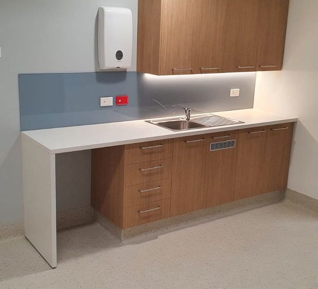 A compact institutional kitchenette or utility kitchen with warm wood-grain cabinetry, a white solid-surface benchtop, stainless-steel sink and drainer, and a muted blue-grey splashback. The space has practical built-in storage, a wall-mounted soap dispenser, power outlets, and a light speckled hard floor finish.