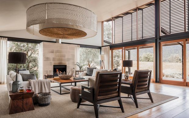 A warm, refined contemporary living room with a large stone fireplace, low-profile seating, dark timber lounge chairs, and expansive timber-framed glazing. Horizontal louvres above the windows provide sun control, while a large woven pendant light and textured neutral furnishings add natural character.