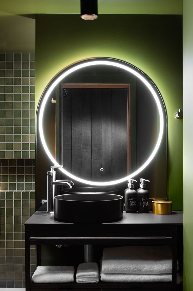 A dramatic, compact bathroom vanity with a large round illuminated mirror, black vessel basin, chrome tapware, dark timber vanity, and deep green walls. Green square tiled wall sections and a black timber door are visible, creating a moody, high-contrast interior.
