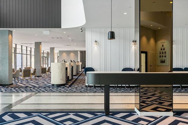 A contemporary hotel or commercial lobby with a long reception or concierge desk in the foreground, sculptural pendant lighting, fluted white wall panelling, dark feature cladding, stone columns, and patterned geometric carpet and flooring. The open-plan space includes lounge seating, decorative screens, and illuminated display niches.