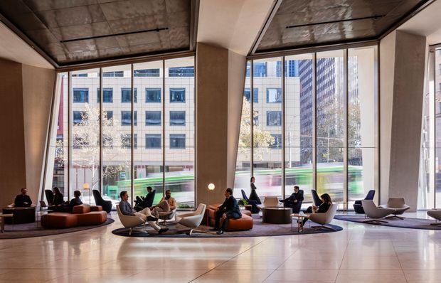 A spacious contemporary commercial lobby or lounge with floor-to-ceiling windows overlooking an urban streetscape. The interior features polished large-format flooring, warm neutral wall surfaces, sculptural lounge seating arranged in small groups, and a reflective metallic ceiling with linear lighting. Several people occupy the seating areas, while a blurred green vehicle is visible outside.