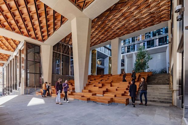 A spacious contemporary covered public atrium or lobby with dramatic exposed timber lattice ceilings, large concrete structural columns, extensive glazing, and stepped timber seating integrated into a broad staircase. People occupy the seating and circulation areas, while modern multi-storey buildings are visible beyond.