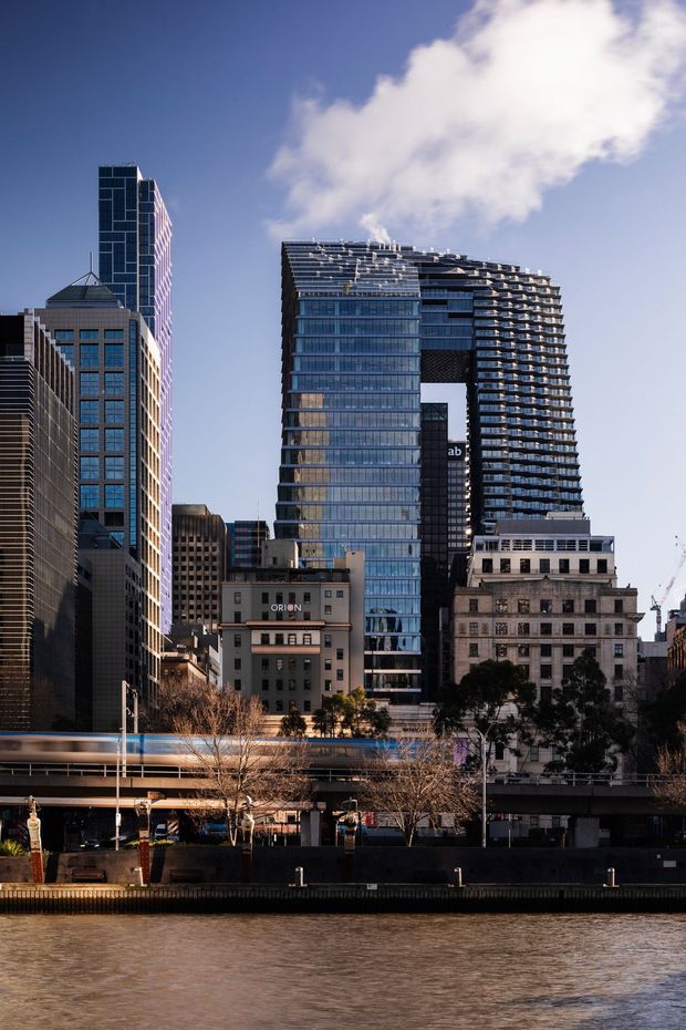 Urban waterfront cityscape showing a cluster of high-rise commercial and mixed-use buildings, including a prominent contemporary tower with extensive glass curtain walling, projecting balconies, and a large elevated void. Older mid-rise masonry buildings and elevated transport infrastructure line the river in the foreground.