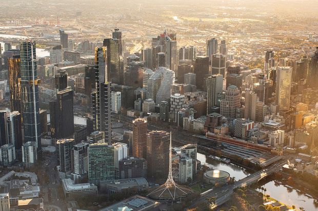 Aerial panoramic view of a dense modern city centre with numerous high-rise towers, office buildings, residential skyscrapers, major rail infrastructure, roads, a stadium-like circular structure, and a waterfront or landscaped precinct. The scene is photographed in warm late-afternoon light and is dominated by urban architecture and metropolitan density.