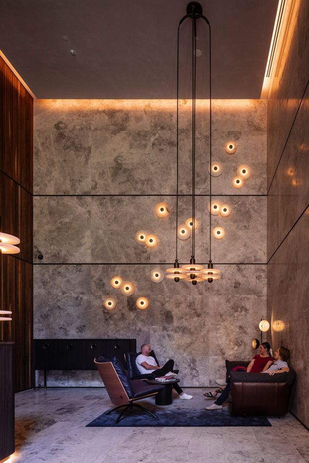 A dramatic double-height lounge or living room with large-format grey stone wall and floor surfaces, dark timber wall panelling, and warm concealed cove lighting. A sculptural linear pendant hangs over a seating area with leather sofas, lounge chairs, and a dark rug. Decorative wall lights create a constellation-like pattern across the stone feature wall, giving the space a luxurious, cinematic atmosphere.