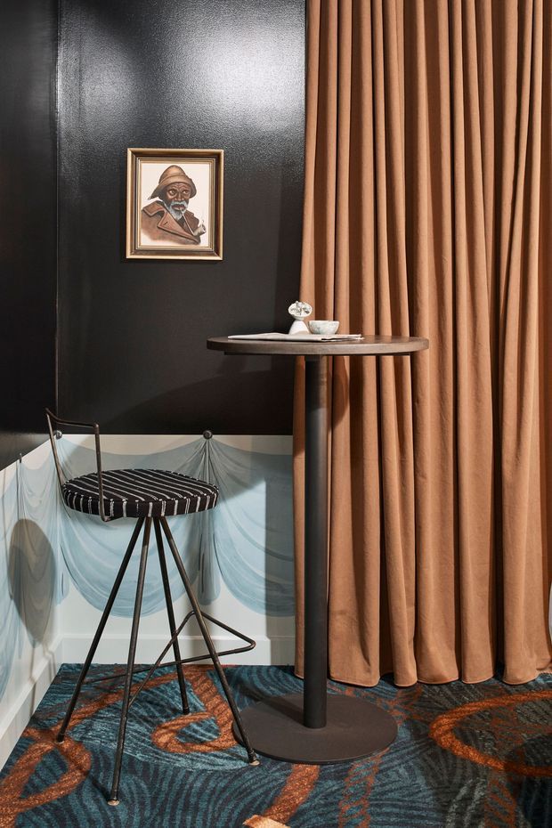 A stylised interior corner featuring a tall round dark wood or stone pedestal table beside a black-and-white striped bar stool. A black textured wall is decorated with a small framed portrait, while pale blue sculptural wall panelling runs along the lower portion. Full-height pleated tan curtains occupy the right side, and a richly patterned blue, rust and charcoal carpet covers the floor. The scene has a theatrical, retro-inspired hospitality or lounge aesthetic.