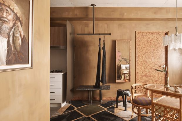 Warm, richly layered interior showing a compact open-plan living and dining area with timber-panelled walls and ceiling beams, a dark geometric rug, woven cane furniture, a small round dining table, artwork, mirror, sculptural pendant lighting, and a partially visible kitchen. The space has an eclectic, refined character with strong natural materials and dark contrasting details.