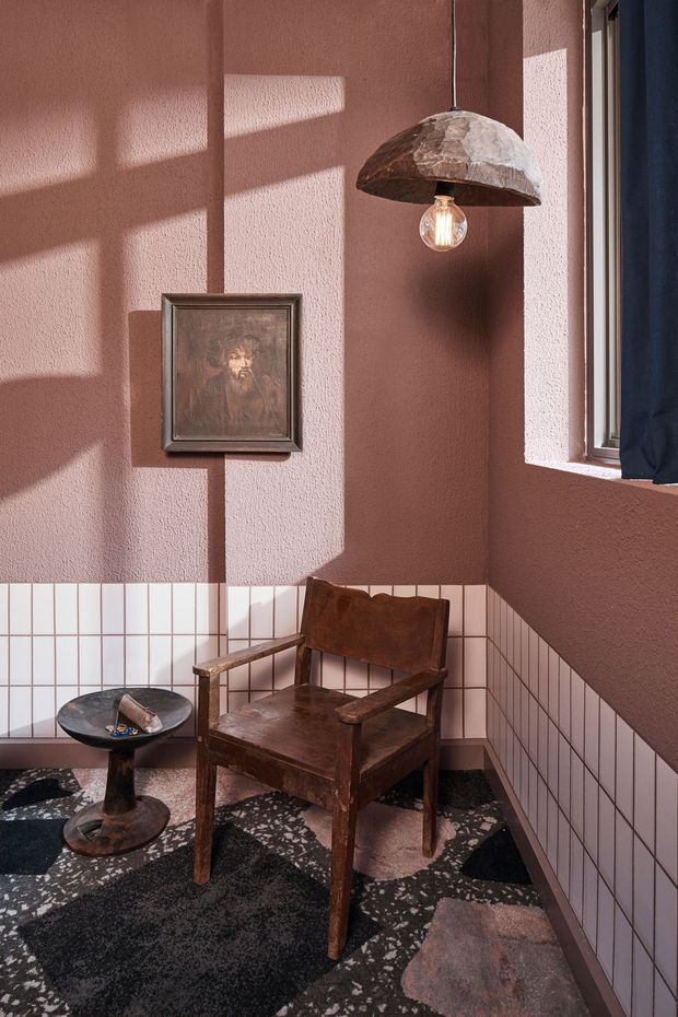 A compact, stylised interior corner with dusty terracotta textured walls, white square tiled wainscoting, and a dark terrazzo floor. A vintage dark timber and brown leather armchair sits beside a small black pedestal side table. A framed artwork hangs on the wall, while a sculptural rough-textured pendant light and strong sunlight create graphic shadows. A window with dark blue curtains is visible at right.