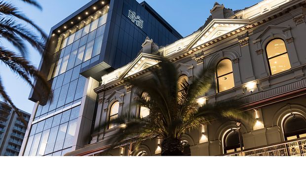 Exterior architectural photograph of a restored ornate historic building facade with classical detailing, arched windows, decorative cornices, pilasters and a balustrade, juxtaposed against a taller contemporary glass-and-dark-metal office building. Palm trees frame the urban streetscape, and warm architectural lighting illuminates the facade at dusk.