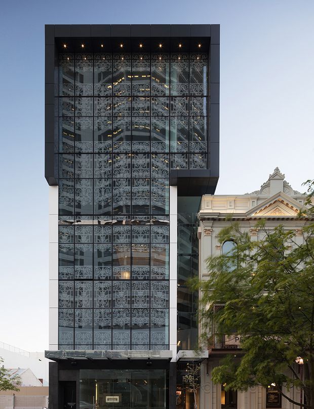Street-level exterior view of a contemporary multi-storey commercial building with a tall dark-framed glazed facade. Decorative patterned screens or graphics are visible behind the curtain wall, with recessed lighting at the upper level and a projecting dark canopy near the base. The modern building is closely attached to an ornate historic classical facade, creating a contrasting heritage-and-contemporary streetscape. Trees partially obscure the lower right portion.