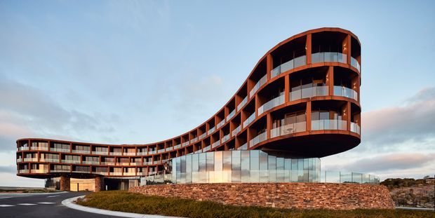 Exterior architectural photograph of a large contemporary multi-storey building with a sweeping, curved form and prominent rounded end volume. The structure features extensive floor-to-ceiling glazing, continuous balconies with transparent balustrades, warm rust-coloured vertical framing, and a substantial stone-clad retaining wall at the base. It is set on an elevated landscaped site beneath an open sky.