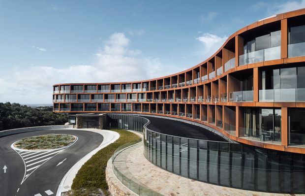 Exterior architectural photograph of a large contemporary multi-storey building with a sweeping curved plan and warm orange-brown facade framing extensive floor-to-ceiling glazing. A curved driveway and pedestrian paving lead around the building, with landscaped planting and distant tree-covered surroundings visible beyond.